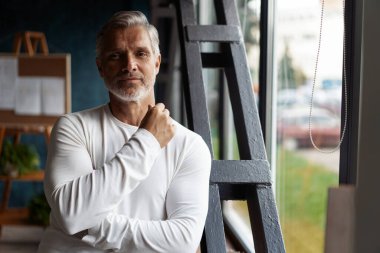 Gülümseyen, sıradan, gri saçlı, olgun bir adam pencerenin yanında kameraya bakıyor..