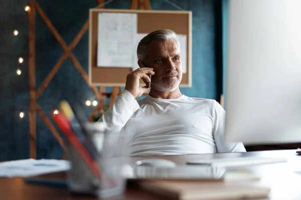 Grijsharige volwassen zakenman aan tafel, werkend op de computer, pratend op de smartphone. Telefoongesprekken. — Stockfoto