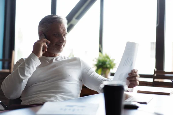 Grijsharige volwassen zakenman aan tafel, werkend op de computer, pratend op de smartphone. Telefoongesprekken. — Stockfoto