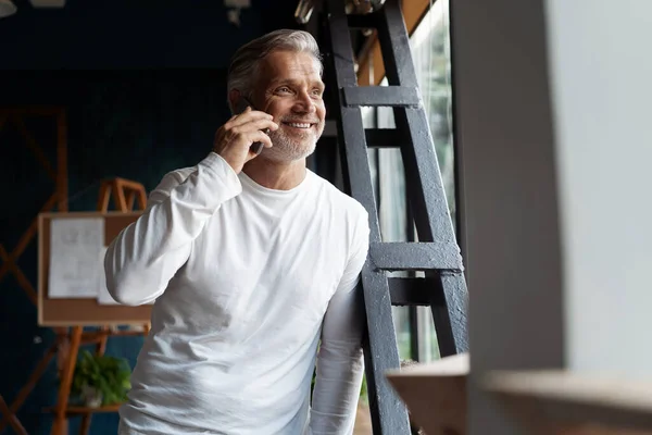 Casual Grey-haired Mature affärsman talar på en mobiltelefon står vid fönstret. — Stockfoto