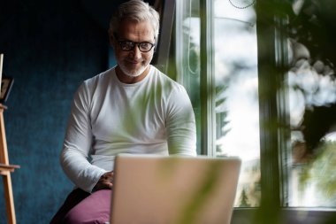 Gri saçlı, güneş gözlüklü, ofiste ya da pencerenin yanındaki kafede çalışırken dizüstü bilgisayarda çalışan yetişkin bir adam.