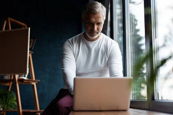 Casual de cabelos grisalhos Maduro homem em óculos trabalhando no laptop enquanto sentado no escritório ou café perto da janela — Fotografia de Stock