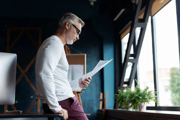 Casual grijs-harige volwassen man lezen van papier in zijn kantoor. — Stockfoto