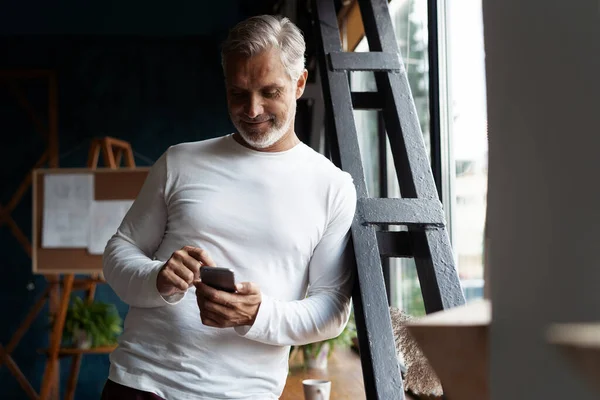 Casual grijs-harige volwassen zakenman met behulp van smartphone staan bij het raam. — Stockfoto