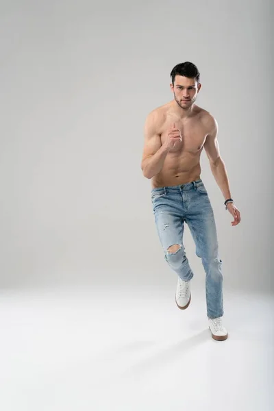 Portrait of a well built shirtless muscular male model running or jumping in air isolated over gray background. — Stock Photo, Image