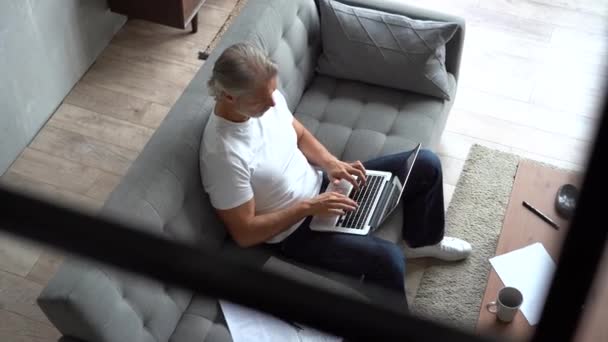 Szary Dojrzały przystojny mężczyzna siedzi na kanapie i pisze na laptopie w domu — Wideo stockowe