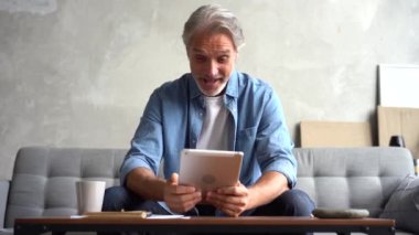 Tablet bilgisayarlı gri saçlı, olgun, mutlu bir adam evde video görüşmesi yapıyor.