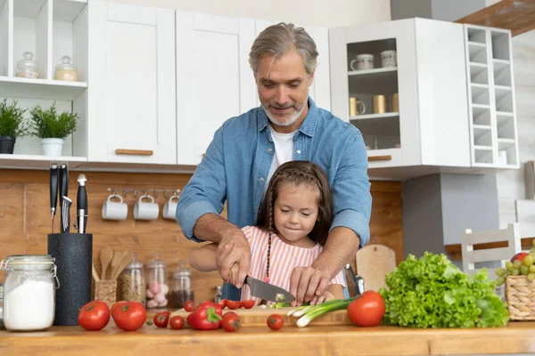 Linda niña y su padre guapo están cortando verduras y sonriendo mientras cocinan en la cocina en casa. — Foto de Stock