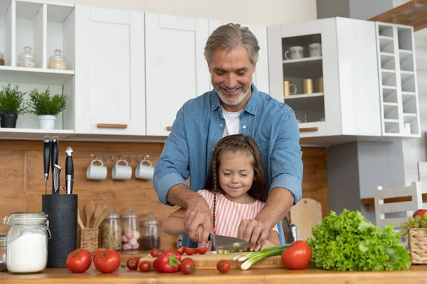 Linda niña y su padre guapo están cortando verduras y sonriendo mientras cocinan en la cocina en casa. — Foto de Stock