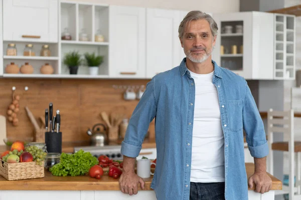 Ősz hajú, jóképű fehér férfi áll a világos konyhában. — Stock Fotó