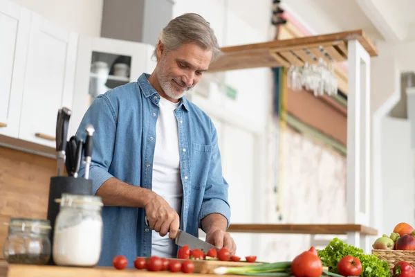 Hombre maduro guapo de pelo gris preparando comida deliciosa y saludable en la cocina casera. — Foto de Stock