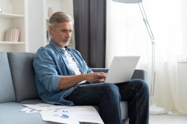 Uzaktan kumanda. Gri saçlı, olgun, yakışıklı, beyaz adam laptopta çalışıyor. Evdeki koltukta oturuyor..