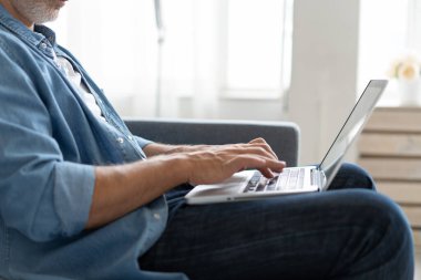 Uzaktan kumanda. Gri saçlı, olgun, yakışıklı, beyaz adam laptopta çalışıyor. Evdeki koltukta oturuyor..