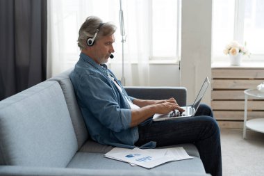 Kulaklıklı Pozitif Adam dizüstü bilgisayar kullanıyor Podcast dinliyor ya da evde koltukta oturan Webinar 'da rol alıyor.