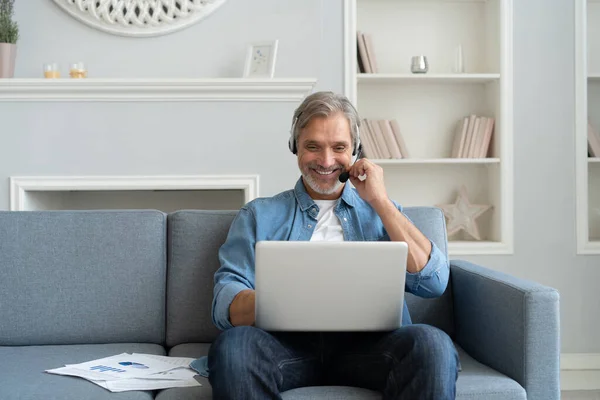 Hombre positivo en auriculares usando el ordenador portátil escuchando Podcast o tomando parte en webinar sentado en el sofá en casa. — Foto de Stock