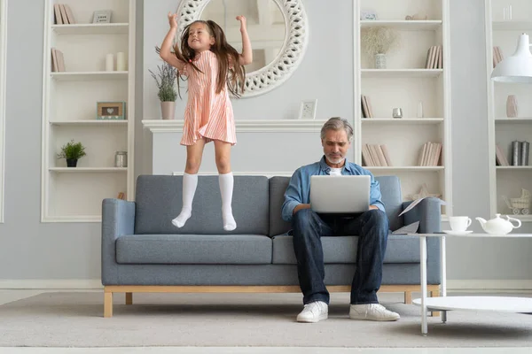 Нет времени для ребенка. Седовласый отец, занятый ноутбуком, работающий дома в интернете, грустная, обиженная дочь, прыгающая рядом — стоковое фото