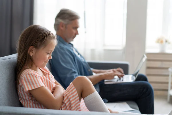 Çocuğa zaman yok. Gri saçlı baba dizüstü bilgisayarla meşgul, evde çalışıyor, üzgün sıkılmış kızı yanında oturuyor. — Stok fotoğraf