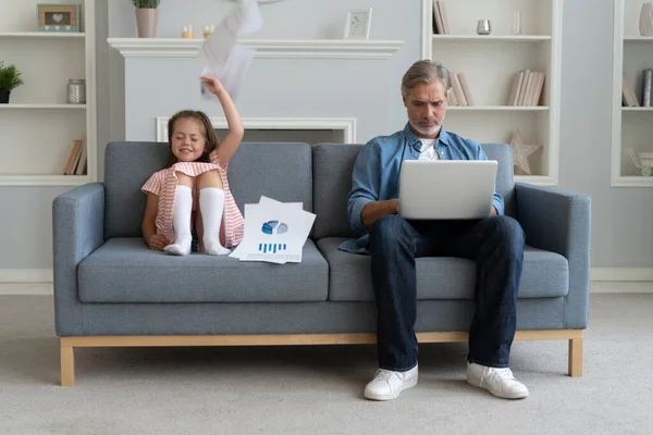Pas le temps pour l'enfant. Papa aux cheveux gris occupé avec un ordinateur portable, travaillant en ligne à la maison, triste ennuyé offensé fille assise à proximité — Photo