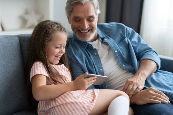 Mignonne fille aider père montrer quelque chose sur smartphone, petite fille intelligente et papa assis sur canapé tenir téléphone mobile — Photo