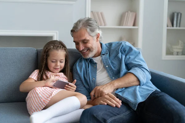 Милая девушка помочь отцу показать что-то на смартфоне, умная маленькая дочь и папа сидеть на диване держать мобильный телефон — стоковое фото