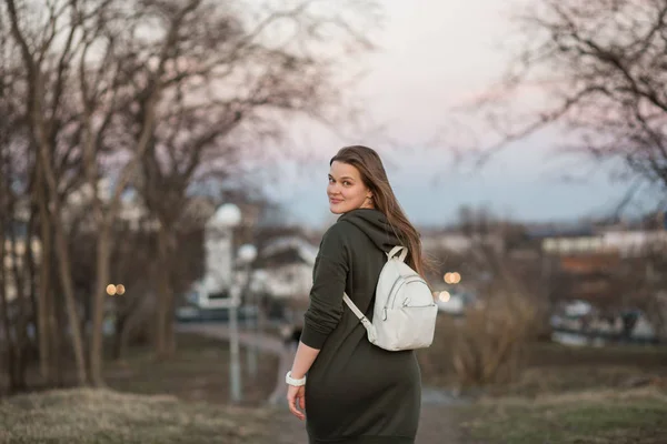 Estilo urbano y concepto de moda. Retrato al aire libre de hermosa elegante modelo femenino joven europeo con el pelo largo marrón con sudadera con capucha de moda y zapatillas blancas relojes, posando en la noche de otoño o — Foto de Stock