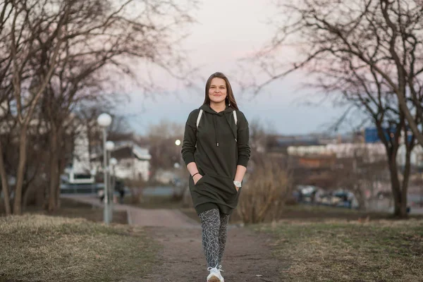 Estilo urbano y concepto de moda. Retrato al aire libre de hermosa elegante modelo femenino joven europeo con el pelo largo marrón con sudadera con capucha de moda y zapatillas blancas relojes, posando en la noche de otoño o — Foto de Stock