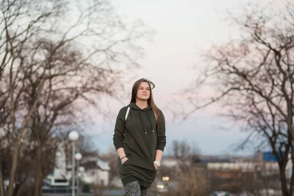 Urbaner Stil und Modekonzept. Outdoor-Porträt der schönen eleganten jungen europäischen Modell mit langen braunen Haaren trägt trendigen Kapuzenpullover und weißen Turnschuhen Uhren, posiert im Herbst abends oder — Stockfoto