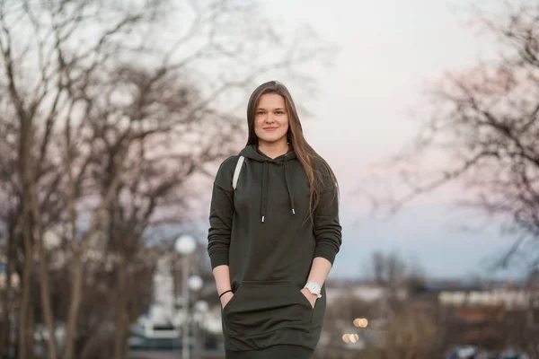 Estilo urbano y concepto de moda. Retrato al aire libre de hermosa elegante modelo femenino joven europeo con el pelo largo marrón con sudadera con capucha de moda y zapatillas blancas relojes, posando en la noche de otoño o — Foto de Stock