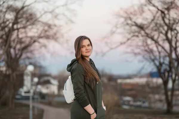 Urban style and fashion concept. Outdoor portrait of beautiful stylish young European female model with long brown hair wearing trendy hoodie and white sneackers watches, posing in evening autumn or — Stock Photo, Image