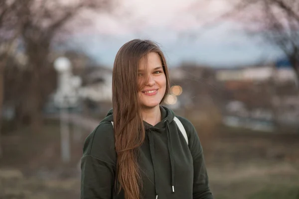 Estilo urbano y concepto de moda. Retrato al aire libre de hermosa elegante modelo femenino joven europeo con el pelo largo marrón con sudadera con capucha de moda y zapatillas blancas relojes, posando en la noche de otoño o — Foto de Stock