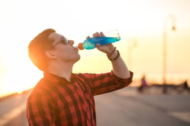 Genç adam mavi izotonik günbatımı gökyüzü arka plan üzerinde sıcak gün sonra şişeden içme. Sağlıklı yaşam ve fitness kavramı