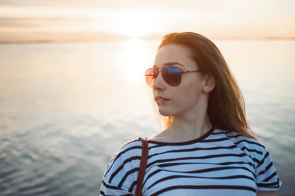 Una joven está mirando el atardecer sobre un mar o un río con hermosos reflejos suaves y soleados en el agua — Foto de Stock