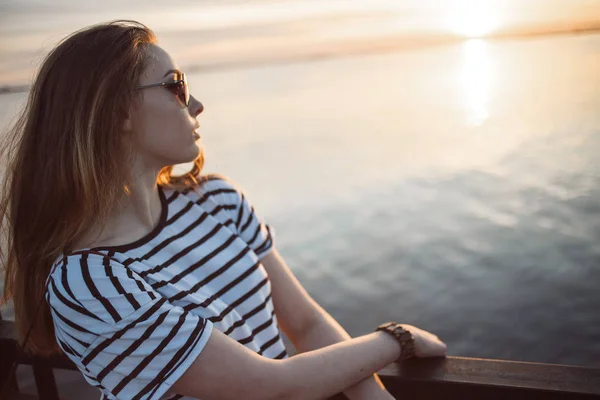 Молодая женщина смотрит на закат над морем или рекой с красивыми мягкими солнечными отражениями в воде — стоковое фото