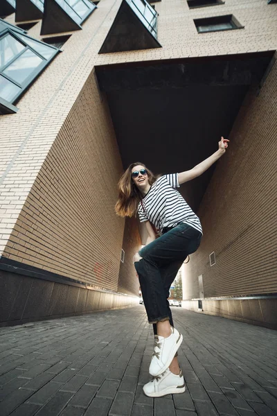 Šťastná dívka tančit a skákat na ulici městské pozadí. Pohled zdola. Hezká žena nosí bílé snickers, tmavé džíny a pruhované tričko. — Stock fotografie