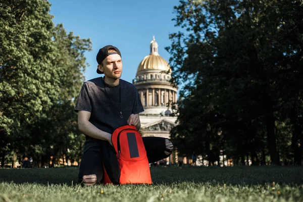 Jugendlicher mann tourist mit rucksack sitzt bei sonnigem sommertag auf einem picknick im gras in der nähe der kathedrale von saint-petersburg — Stockfoto