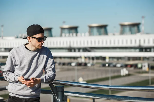 Красивый человек путешественник с телефоном возле аэропорта Пулково в Санкт-Петербурге. Он надел круглые солнцезащитные очки, черную шапочку и серый свитер. — стоковое фото
