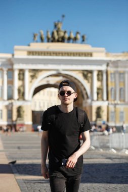 Genç şık Turizm Genel Kurmay Builsing arch palace Square, St. Petersburg yakınındaki yürüyen adam. Riund güneş gözlüğü ile siyah kot pantolon, siyah gömlek, siyah şapkalı giymiş