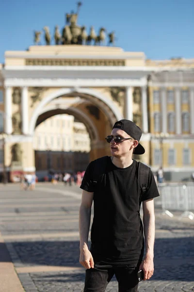Молодой стильный турист, гуляющий возле Генштаба, строит арку на дворцовой площади в Санкт-Петербурге. Он одет в черные джинсы, черную футболку, черную кепку в веселые солнцезащитные очки — стоковое фото