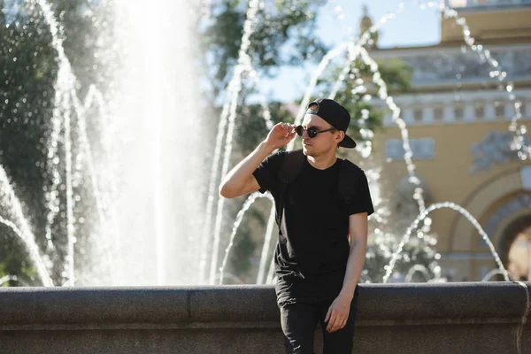 Homem elegante na fonte no dia quente de verão. Usa calças de ganga pretas, t-shirt preta, boné preto com óculos escuros. — Fotografia de Stock