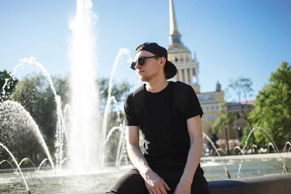 Homem elegante na fonte no dia quente de verão. Usa calças de ganga pretas, t-shirt preta, boné preto com óculos escuros. — Fotografia de Stock