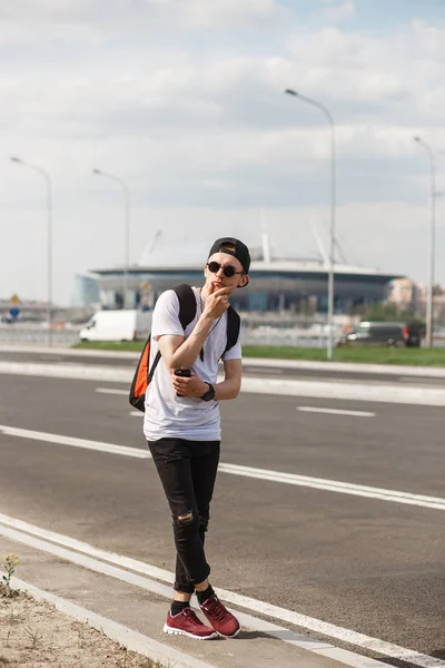 Elegante homem turista na rua no fundo do estádio de futebol borrado. Ele usa calças de ganga pretas, t-shirt preta, boné preto com óculos escuros. Conceito de jogos de futebol, fãs, futebol — Fotografia de Stock