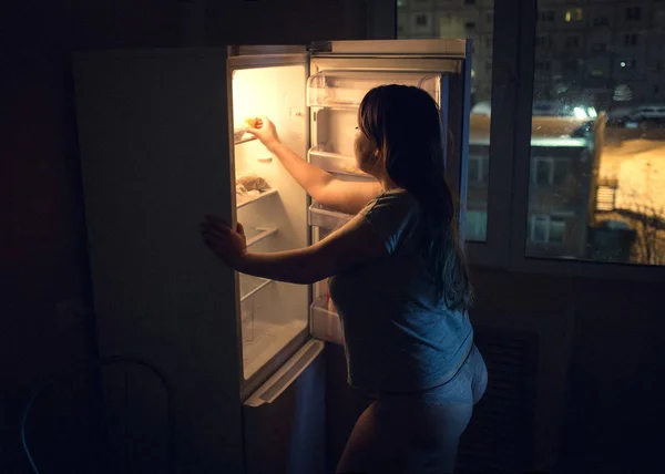 Сексуальная толстушка, жаждущая и ищущая еду в холодильнике поздно ночью. Девушка, одетая в футболку и трусики. Аутентичная атмосфера кухни . Стоковое Фото