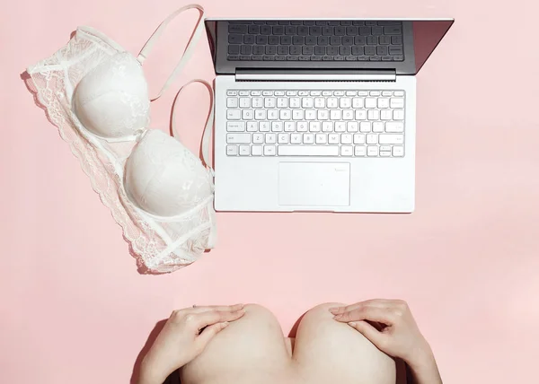 Erotic chat, virtual sex concept. Woman flirting in an online chat. Big female breast covered by hands and white bra thrown near modern laptop, against a pink background. Minimalistic Flat lay, top