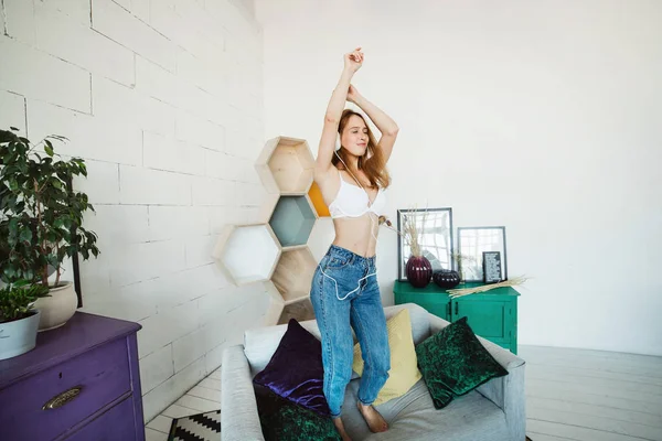 Concept de loisirs, de maison et de technologie - jeune femme heureuse dans un casque blanc écoutant la musique du smartphone et dansant sur le lit à la maison. Elle portait un soutien-gorge blanc et un jean bleu — Photo