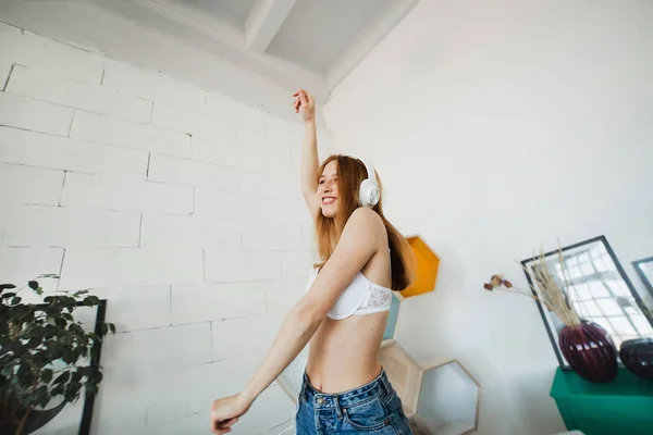 Досуг, дом и технологии концепция - счастливая молодая женщина в белых наушниках, слушающая музыку со смартфона и танцующая на кровати дома. На ней белый лифчик и синие джинсы. — стоковое фото