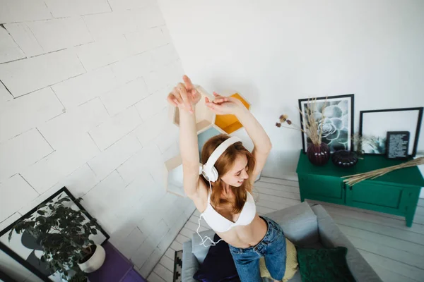 Concepto de ocio, hogar y tecnología: una joven feliz con auriculares blancos escuchando música desde un smartphone y bailando en la cama en casa. Ella llevaba sujetador blanco y vaqueros azules — Foto de Stock