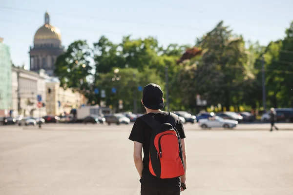 Widok z tyłu młody człowiek stylowy turystycznych chodzenie na Plac Pałacowy w pobliżu Saint Isaacs katedry w Saint-Petersburg. Ubrany w czarne dżinsy, czarny T-shirt, czarny i czerwony plecak — Zdjęcie stockowe