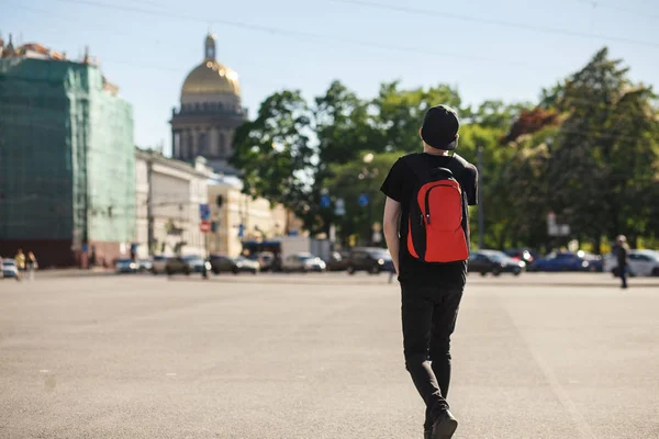 Вид на молодого стильного туриста, гуляющего на Дворцовой площади возле Исаакиевского собора в Санкт-Петербурге. Он одет в черные джинсы, черную футболку, черную кепку и красный рюкзак — стоковое фото