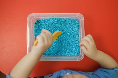 Çocuk duyusal kutusunda mavi renkli pirinç sarı kaşıkla oynayan eller. Babys duyusal eğitim seti