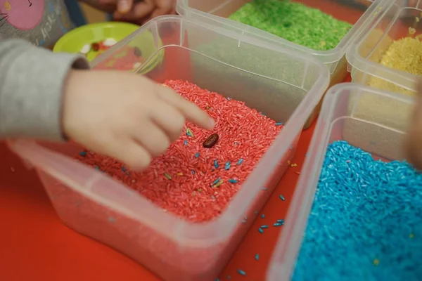 Детские руки играют с цветным рисом в сенсорной коробке. Набор сенсорных учебных материалов для младенцев Лицензионные Стоковые Фото